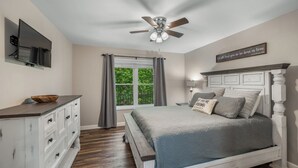 Ein Schlafzimmer mit Bett, Schminktisch, Deckenventilator, Fernseher, Fenster mit Vorhängen und einem motivierenden Zitat an der Wand.
