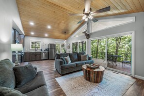 A spacious living area with a wooden ceiling, a large sofa, a coffee table, and a view of the outdoors through large windows. - Mountain Music Romantic Private Brow Cottage with Hot Tub (Fort Payne)
