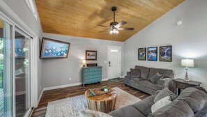 Ein Wohnzimmer mit Holzbalkendecke, einem grauen Sofa, einem Couchtisch und einem an der Wand befestigten Fernseher.