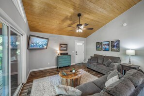 Ein Wohnzimmer mit Holzbalkendecke, einem grauen Sofa, einem Couchtisch und einem an der Wand befestigten Fernseher.
