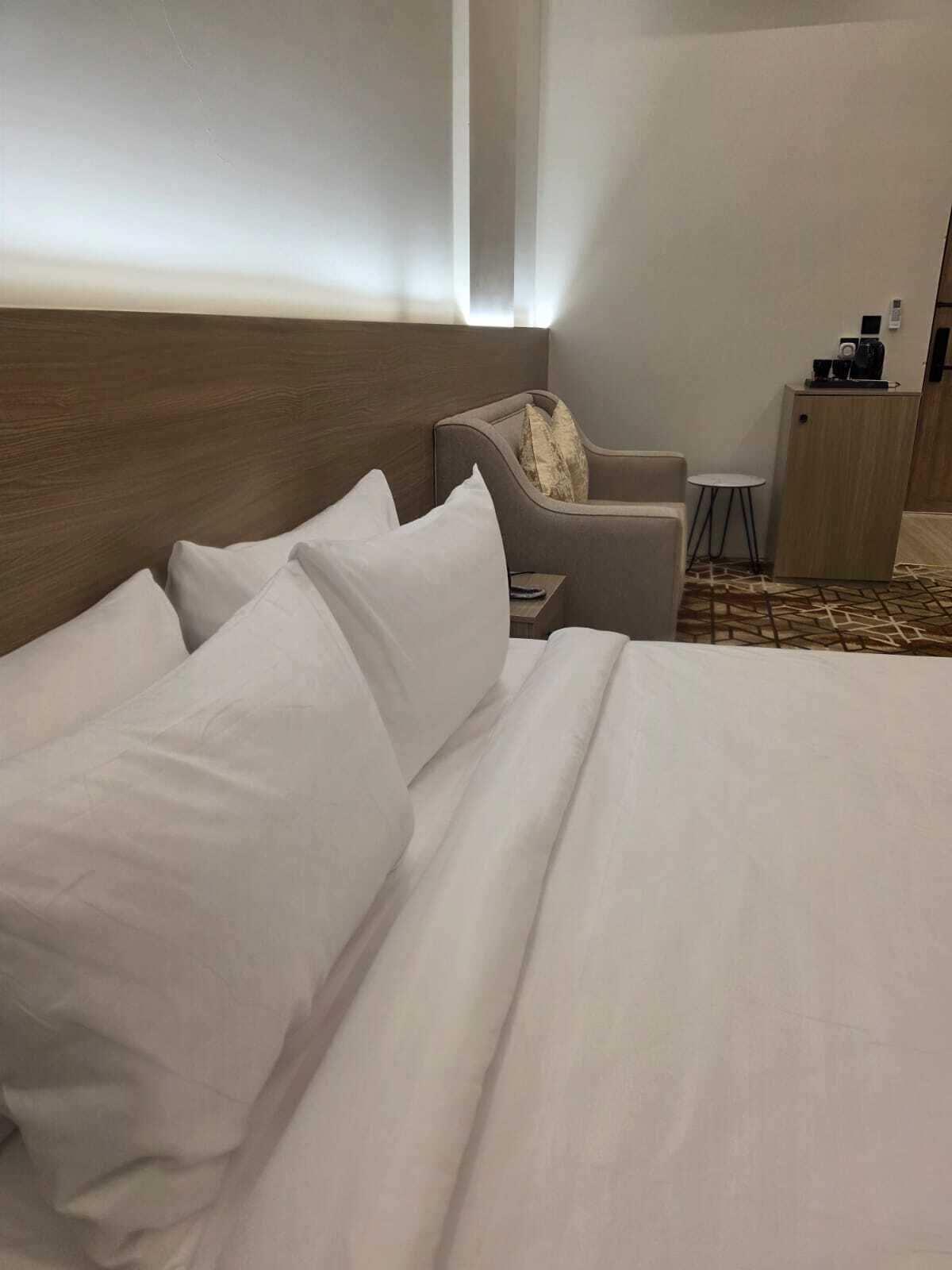 A hotel room with a large bed, a wooden headboard, a small armchair, a side table, and a desk with a chair.