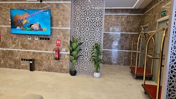 Um hall com televisão, extintor de incêndio, plantas em vasos e um balcão de atendimento.