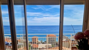 Ein Zimmer mit Meerblick durch große Glasüren, ein Holzboden, ein Tisch mit einer blumigen Tischdecke und ein Sessel.