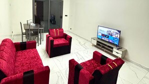 Una sala de estar con un televisor de pantalla plana, sofás rojos y negros, un comedor con una mesa y sillas, y una puerta que da a otra habitación.