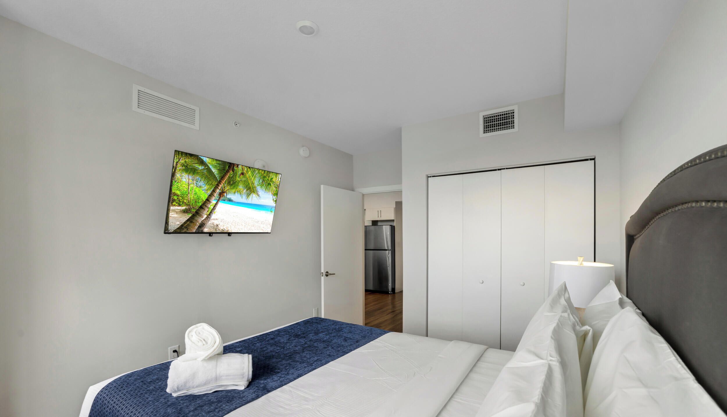 A hotel room with a bed, a television displaying a beach scene, a wardrobe, and a kitchenette.