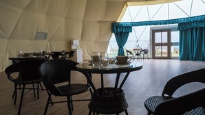 Un'area pranzo con tende, tavoli e sedie rotonde, e vista all'esterno tramite ampie finestre, con una porta che dà sull'esterno.