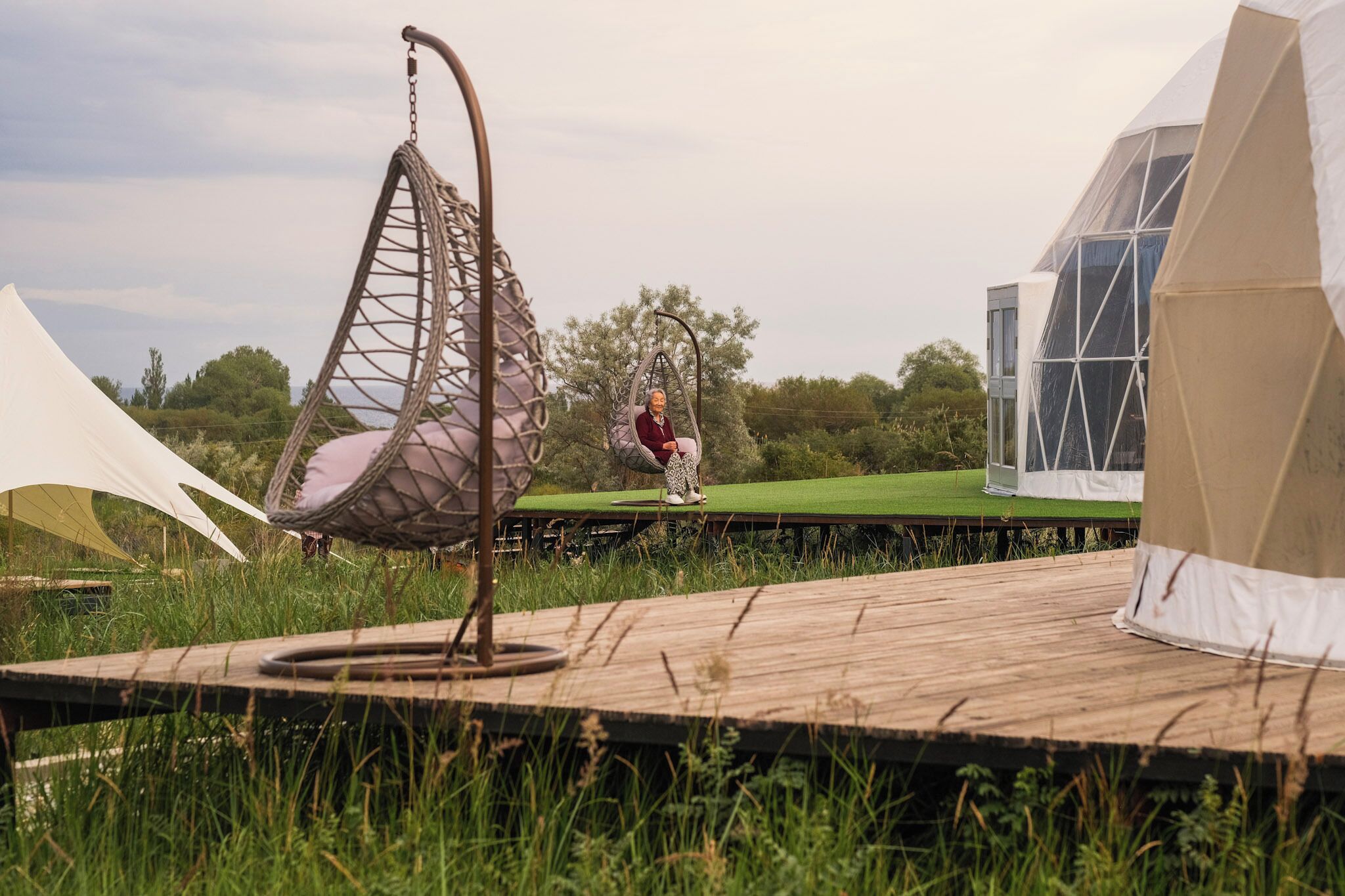 A person sitting on a hanging chair in a grassy outdoor area with tents and a wooden deck.