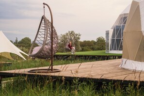 A person sitting on a hanging chair in a grassy outdoor area with tents and a wooden deck. - Nomad Space (Bar-Bulak)