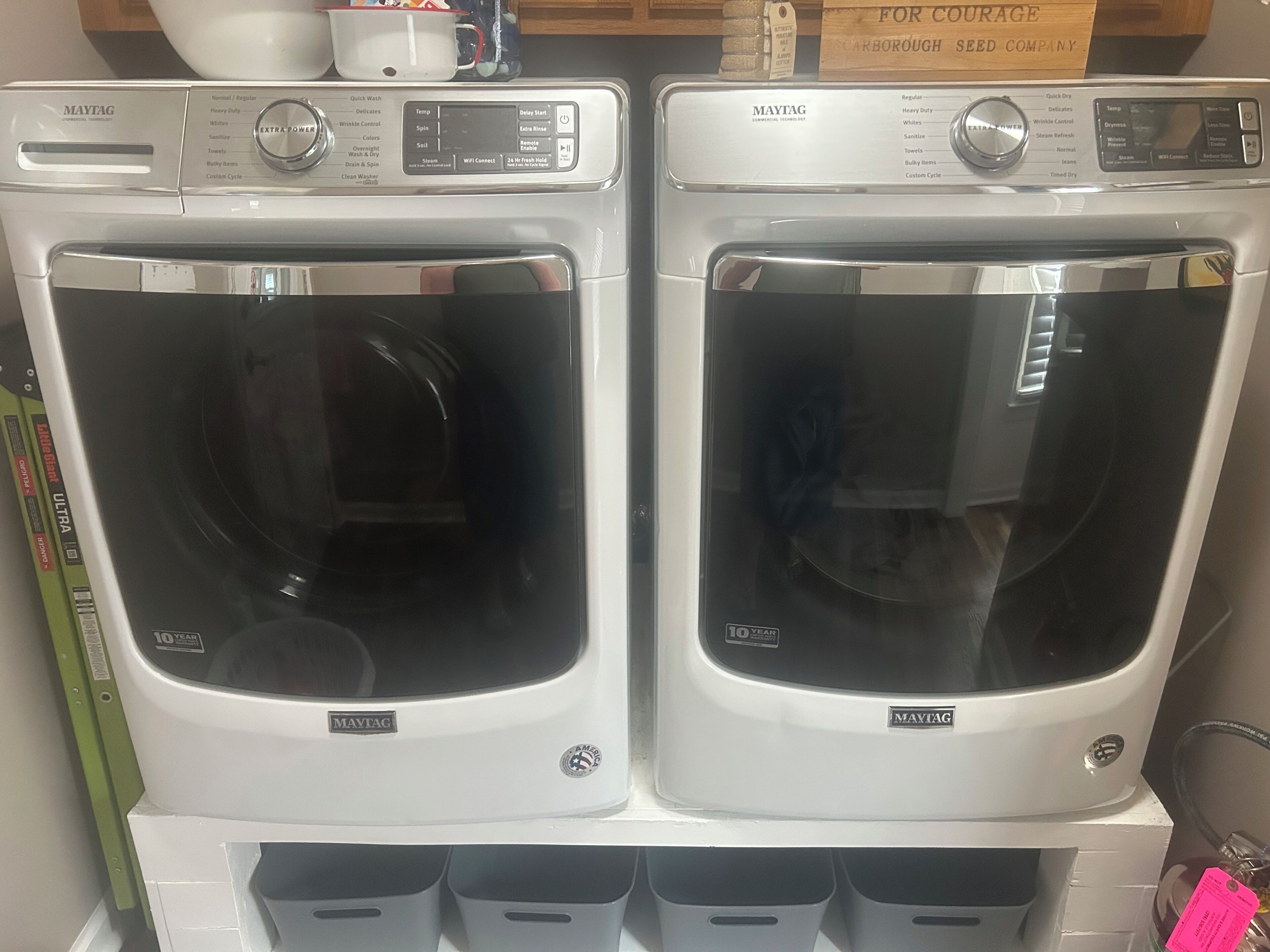 Two Maytag front-loading washing machines with a 10-year warranty label, placed on a white stand with a shelf below.