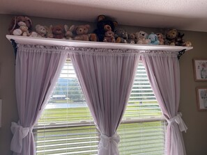 A room with a window featuring a curtain with a bow, a shelf above the window with stuffed animals, and a wall with framed pictures.