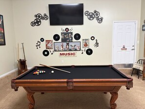 A pool table with a cue stick, surrounded by wall decorations including a television, framed pictures, and vinyl records. - Magnolia House In the Cove. Sweet Southern hospitality awaits you here. (Owens Cross Roads)