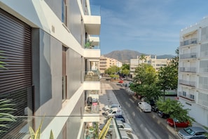 Ein modernes Gebäude mit Balkon, Straßenansicht, parkenden Autos und einem Berg im Hintergrund.