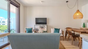 A modern living room with a sofa, a flat-screen TV, a dining table with chairs, and a large window offering a view of buildings and greenery. - Zaga3c - New Modern Apartment in Central Estepona (Estepona)