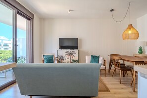 Ein modernes Wohnzimmer mit einer Couch, einem Flachbildfernseher, einem Esstisch mit Stühlen und einem großen Fenster mit Blick auf Gebäude und Grünflächen.