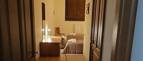 Un pasillo estrecho con una cama y mesita de noche a lo lejos, un banco en el centro, y puertas de madera a ambos lados.