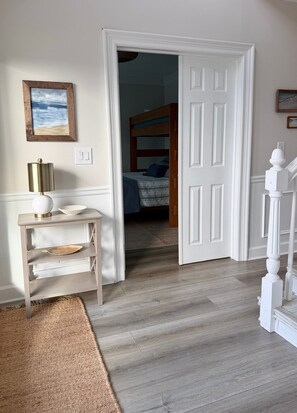 A room with a wooden floor, a white door, a bed with a blue bedspread, a small table with a lamp, and a picture on the wall. - A Rare Bird:Spacious 5-bedroom, beachfront home in welcoming Gulf Shores with AC (Gulf Shores)
