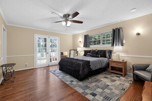 Ein Schlafzimmer mit Bett, Deckenventilator, Fenster mit Vorhängen, Nachttisch, Stuhl und kleinem Tisch.