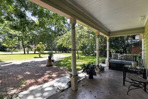 Eine überdachte Veranda mit einem Ziegelweg, der zu einer Rasenfläche führt, einem Sitzbereich mit einer Couch und Blumentöpfen.
