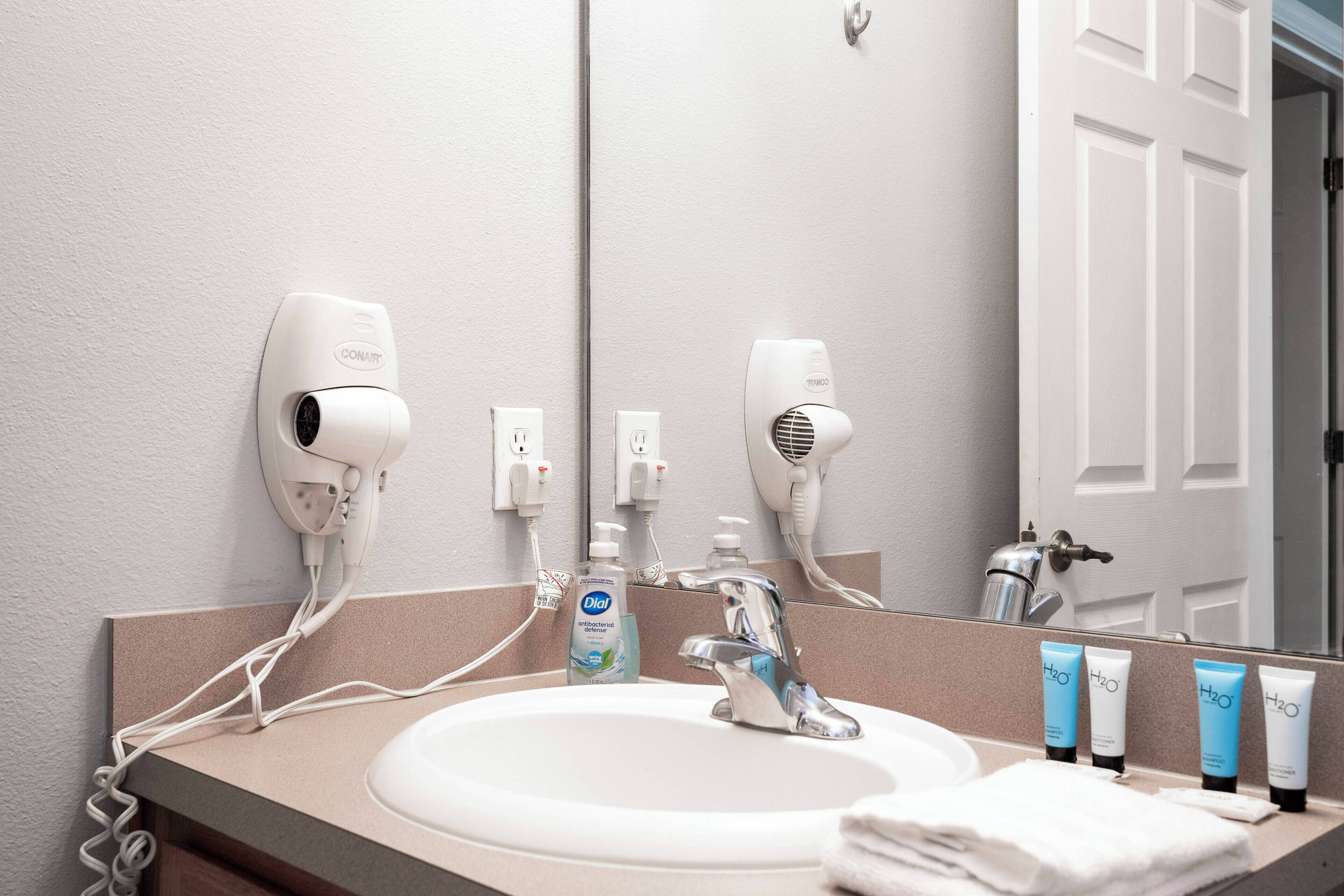 Ein Badezimmer mit Waschbecken, zwei Haartrocknern, einem Spiegel und Pflegeprodukten.
