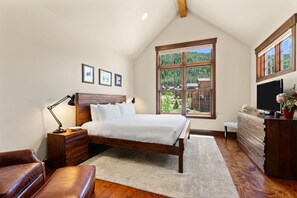 Een slaapkamer met een houten bed, nachtkastje, dressoir, televisie en een groot raam met uitzicht op bomen en gebouwen.