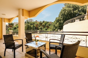 A balcony with wicker chairs, a glass-top table, and a view of a lush garden and buildings. - Quartz 5 Apartment (Cannes)