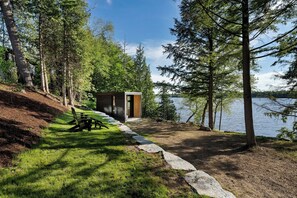 A lakeside cabin with a small deck, a picnic table, and a wooden structure. - Modern Luxury Lakehouse (Stone Mills)