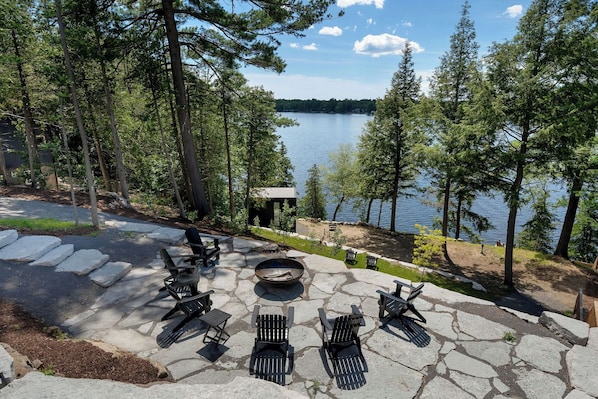 A lakeside patio with stone seating and a fire pit, surrounded by trees and overlooking a lake. - Modern Luxury Lakehouse (Stone Mills)