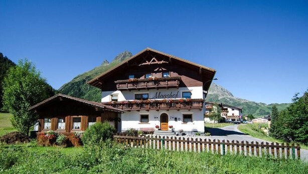 A two-story mountain chalet with a wooden facade, a balcony with flower boxes, and a mountain backdrop. - Maashof (Galtür)