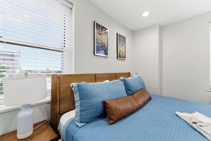 Un dormitorio con una cama, una mesita de noche, un ventanal con persianas y dos cuadros en la pared.