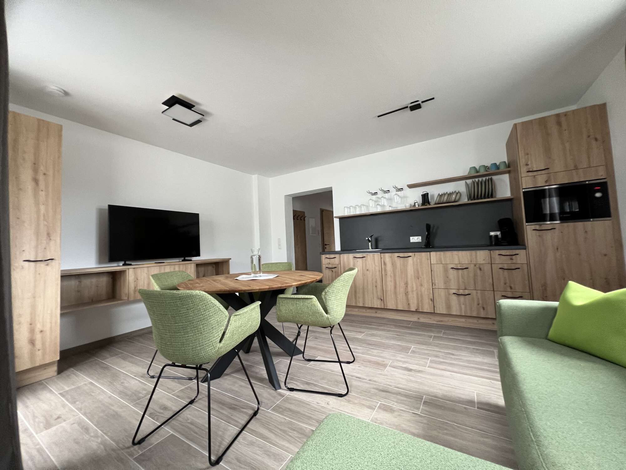 A modern kitchen with wooden cabinets, a dining area with a round table and green chairs, and a flat-screen TV mounted on the wall.