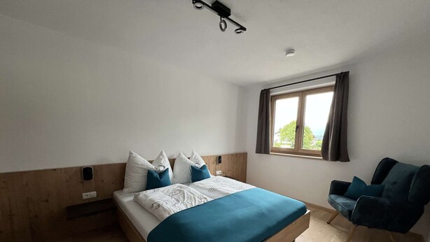 Une chambre à coucher avec un lit en bois, une fenêtre avec des rideaux, une chaise et un luminaire fixé au plafond.
