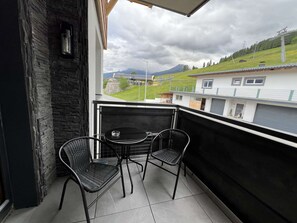Ein Balkon mit zwei Stühlen und einem Tisch, der auf eine grüne Landschaft mit Gebäuden blickt.