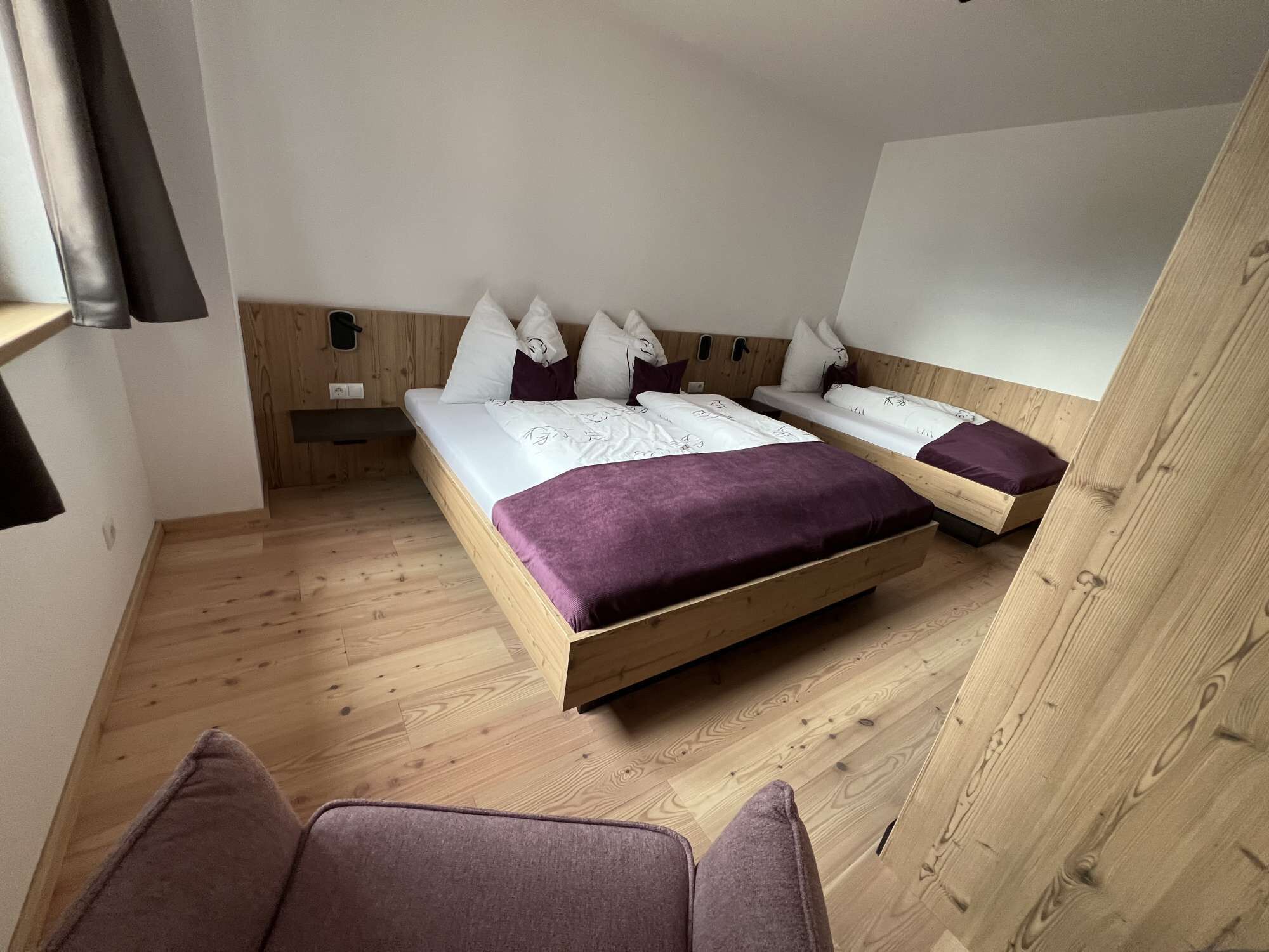 A room with a wooden floor, two beds with white bedding and purple accents, a small bedside table, and a chair.