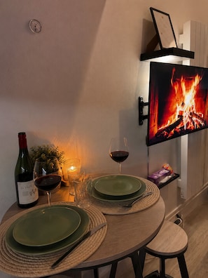 Taula de menjador amb plats, una ampolla de vi, vasos i una espelma. Un televisor de pantalla plana està lluent a la paret, mostrant un foc.
