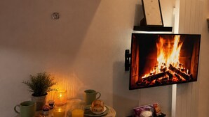 A cozy room with a flat-screen TV displaying a fireplace, a dining table with breakfast items, and a small shelf with a box. - Le Nid Vaillant - Near the Train Station & Free Parking (Tours)