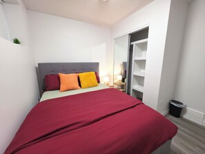 Une chambre à coucher avec un lit, une tête de lit rembourrée de couleur grise, une couvre-lit rouge, deux oreillers, une table de chevet avec une lampe et une armoire dont les portes sont ouvertes.