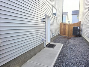 A residential building with a gated backyard, a small concrete pathway, and a visible air conditioning unit. - Cozy 2 Bedroom Legal Suite/Sleeps 4 (Edmonton)