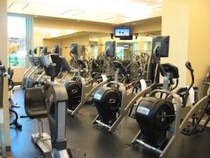Ein Fitnessstudio mit verschiedenen Cardiogeräten, Spiegeln und einem Bildschirm.