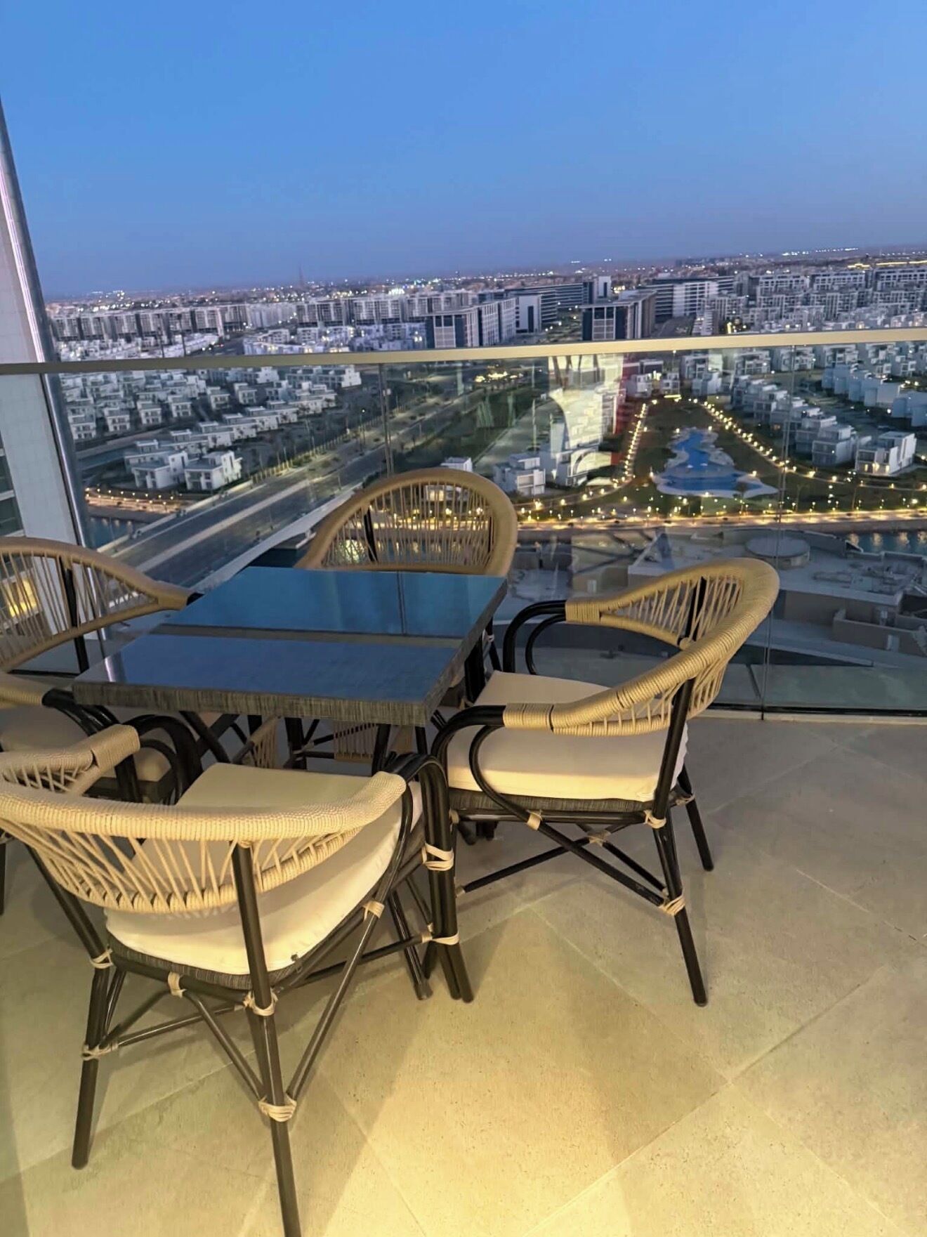 Een balkon van een hoog gebouw met een glazen tafel en rieten stoelen, met uitzicht op een stadsbeeld in de schemering.