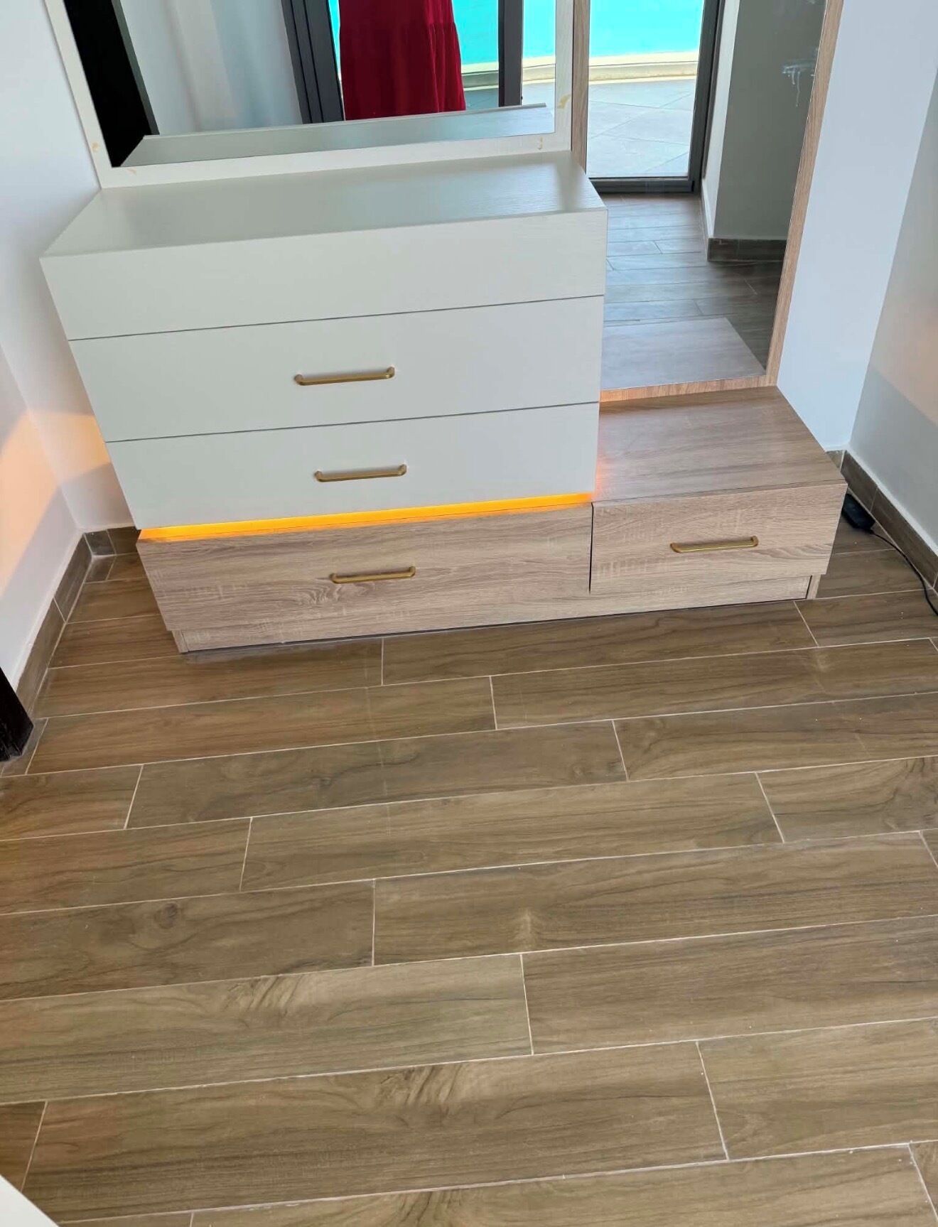 Um quarto com piso de madeira, um armário branco com gavetas e uma peça de roupa vermelha ao fundo.