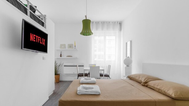 Moderni hotellihuone, jossa on sänky, työpöytä, tuoli, televisio, jossa näkyy Netflix, ja vihreä riippuvalaisin.