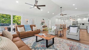 Ein geräumiges Wohnzimmer mit einer braunen Ledersofa, einem Couchtisch und einem gemusterten Teppich. Es gibt Deckenventilatoren, eine Küchenzeile mit weißen Schränken und Blick ins Grüne.