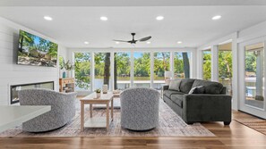 A modern living room with a fireplace, a ceiling fan, a large window with a lake view, and a grey sofa. - The Ozark Acre 6bdr Lakefront (Sunrise Beach)