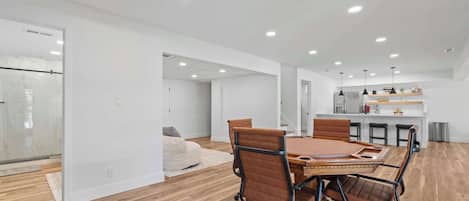 A modern interior with a dining area featuring a wooden table and leather chairs, a kitchen with pendant lighting, and a hallway leading to a bathroom.