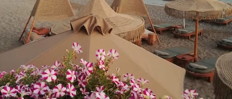 A beachside lounge area with thatched umbrellas and lounge chairs, overlooking a calm sea and distant hills.