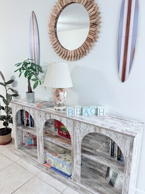 A white wall with a round mirror, two surfboards, a potted plant, a lamp, and a bookshelf with the word 'BEACH' on it. - Sun&Sand Oasis - Beach Front - 2/2 Condo - Next to Heated Pool - Ground Floor (St. Augustine)
