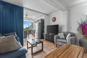 Ein modernes Wohnzimmer mit einer Couch, einem Couchtisch und einem Fernseher. Es gibt eine Küchenzeile mit Schränken und einem Kühlschrank.
