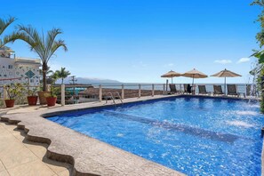 Ein Swimmingpool mit Liegestühlen und Sonnenschirmen, umgeben von Blumentöpfen und freiem Blick auf das Meer.