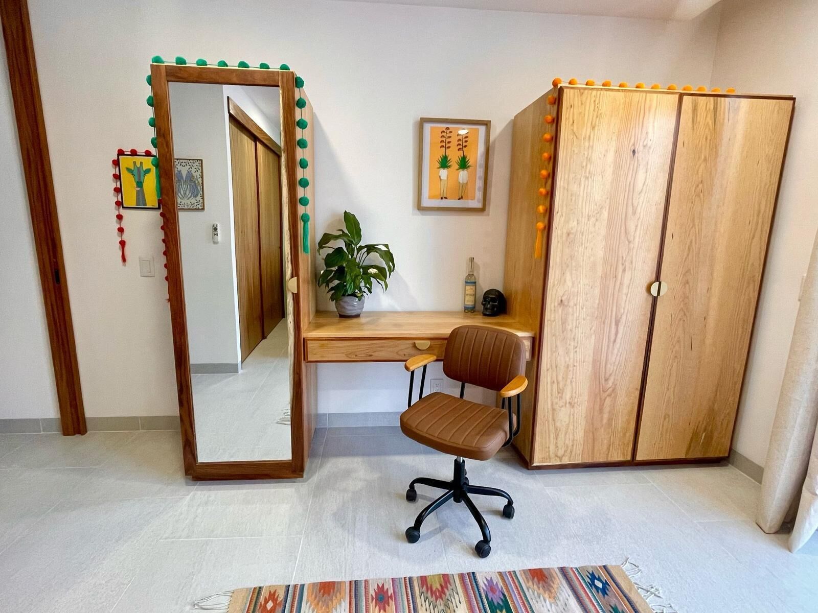 Una habitación con un escritorio de madera, una silla, un armario, un espejo, una planta y una alfombra.
