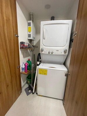 A compact laundry room with a washing machine and dryer, a small shelf with cleaning supplies, and a vent on the wall. - Casa Lluvia Retreat • Rooftop Infinity Pool (Puerto Vallarta)